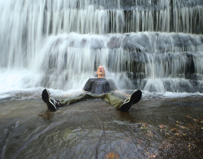 A person is sitting in a shallow pool of water directly under a cascading waterfall. The water rushes over their head and shoulders as they recline with their legs extended and feet pointing upwards. They are wearing dark clothing and shoes, and the surrounding environment appears to be a natural, rocky area with foliage.